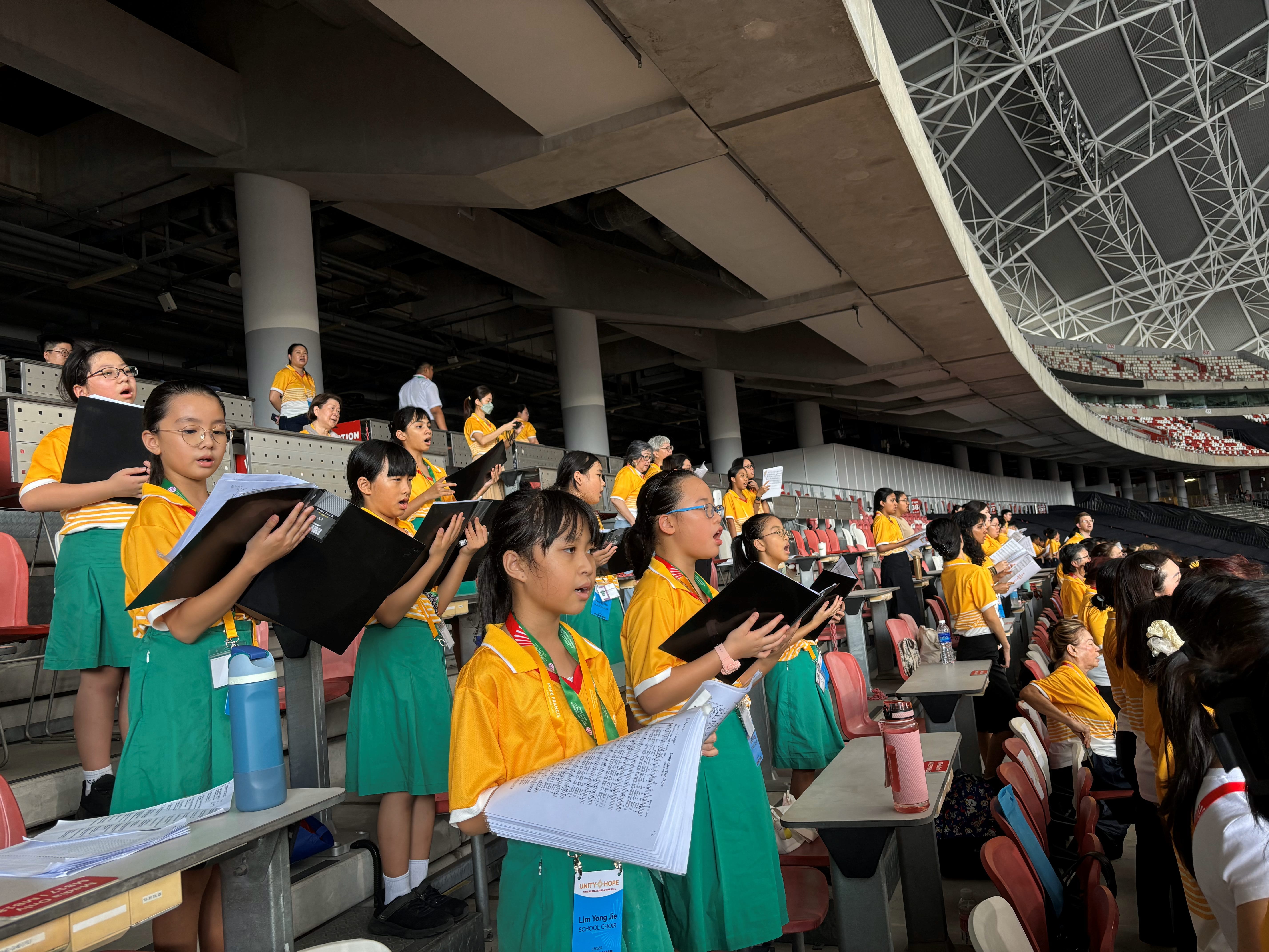 Children in yellow and green uniforms hold sheet music folders and sing in stadium stands.